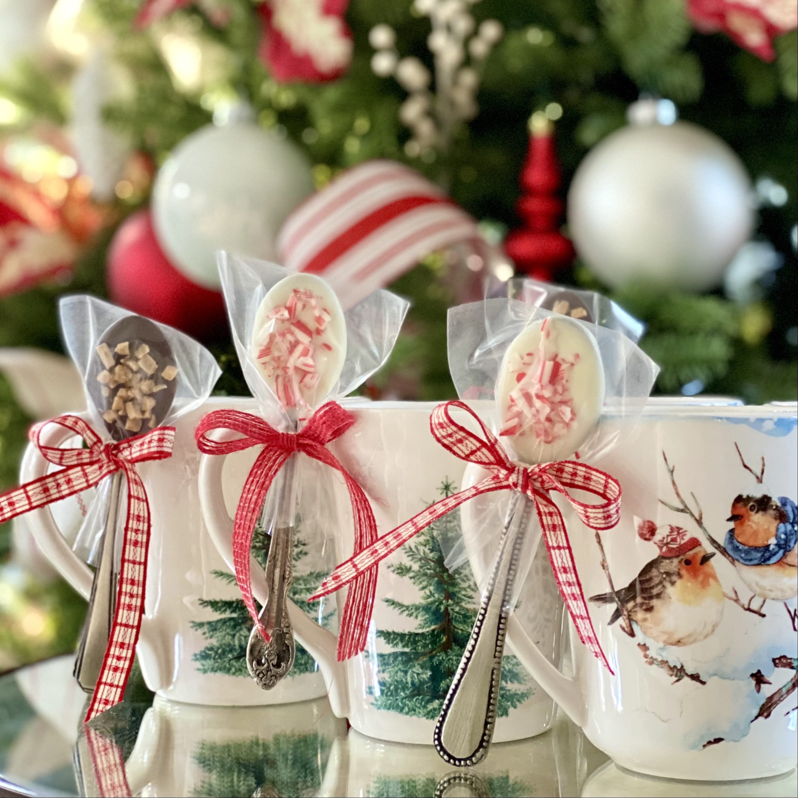 Chocolate dipped spoons tied to Christmas mugs in front of a Christmas tree.