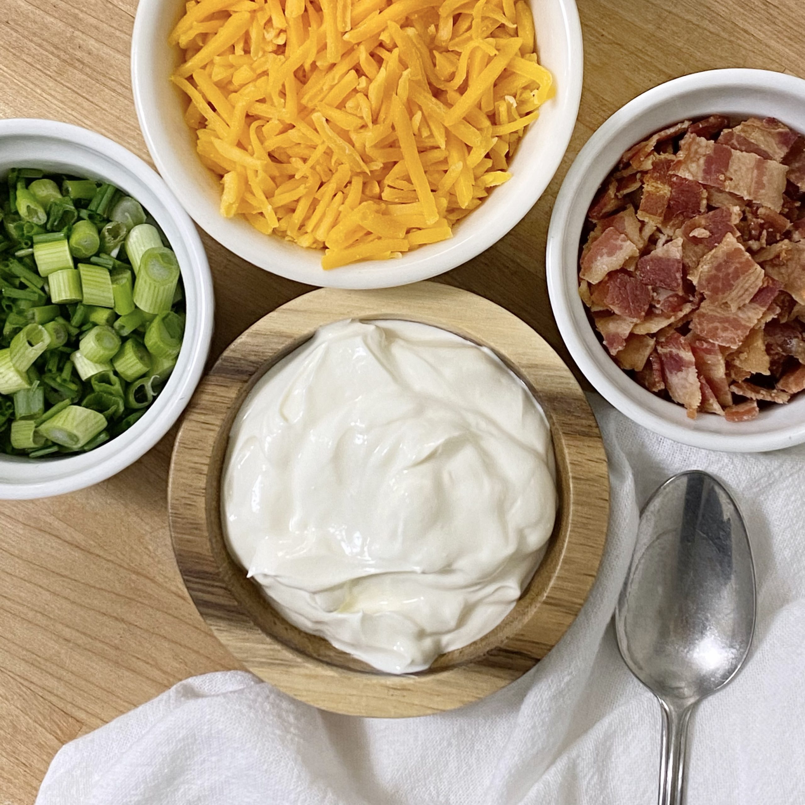 Loaded potato soup toppings including cheddar cheese, crumbled bacon, sour cream, chives and onions with a spoon.