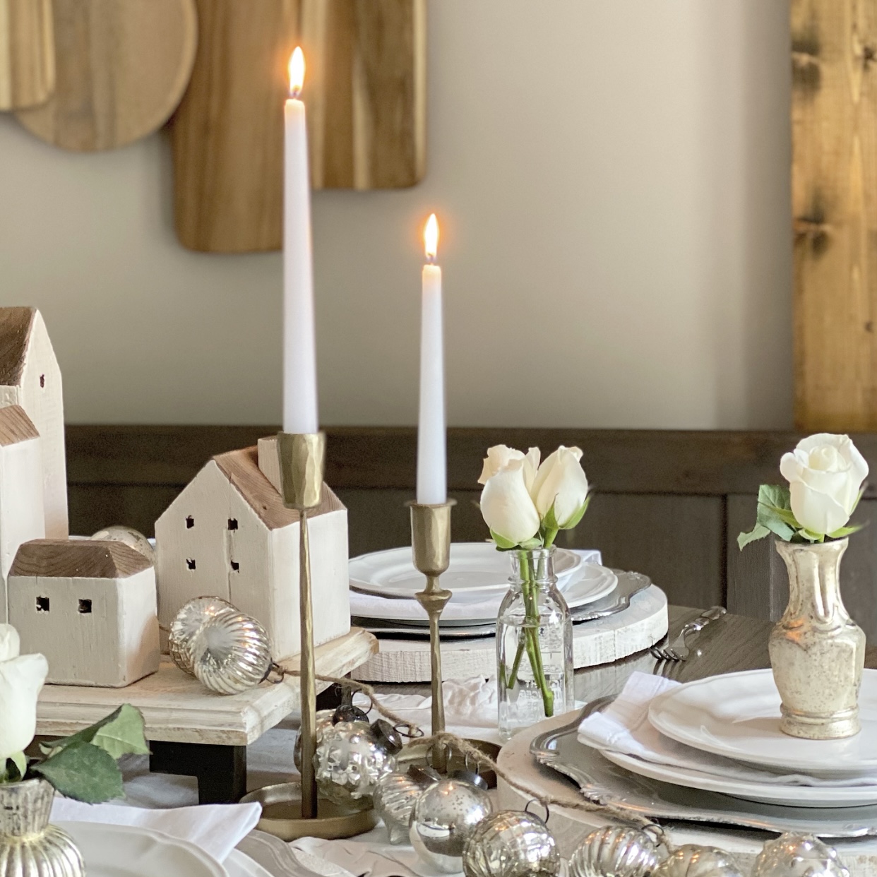 A neutral holiday tablescape with taper candles burning in brass candlesticks, white roses in mercury glass bud vases on each place setting, and reclaimed wood houses in the center of the table.