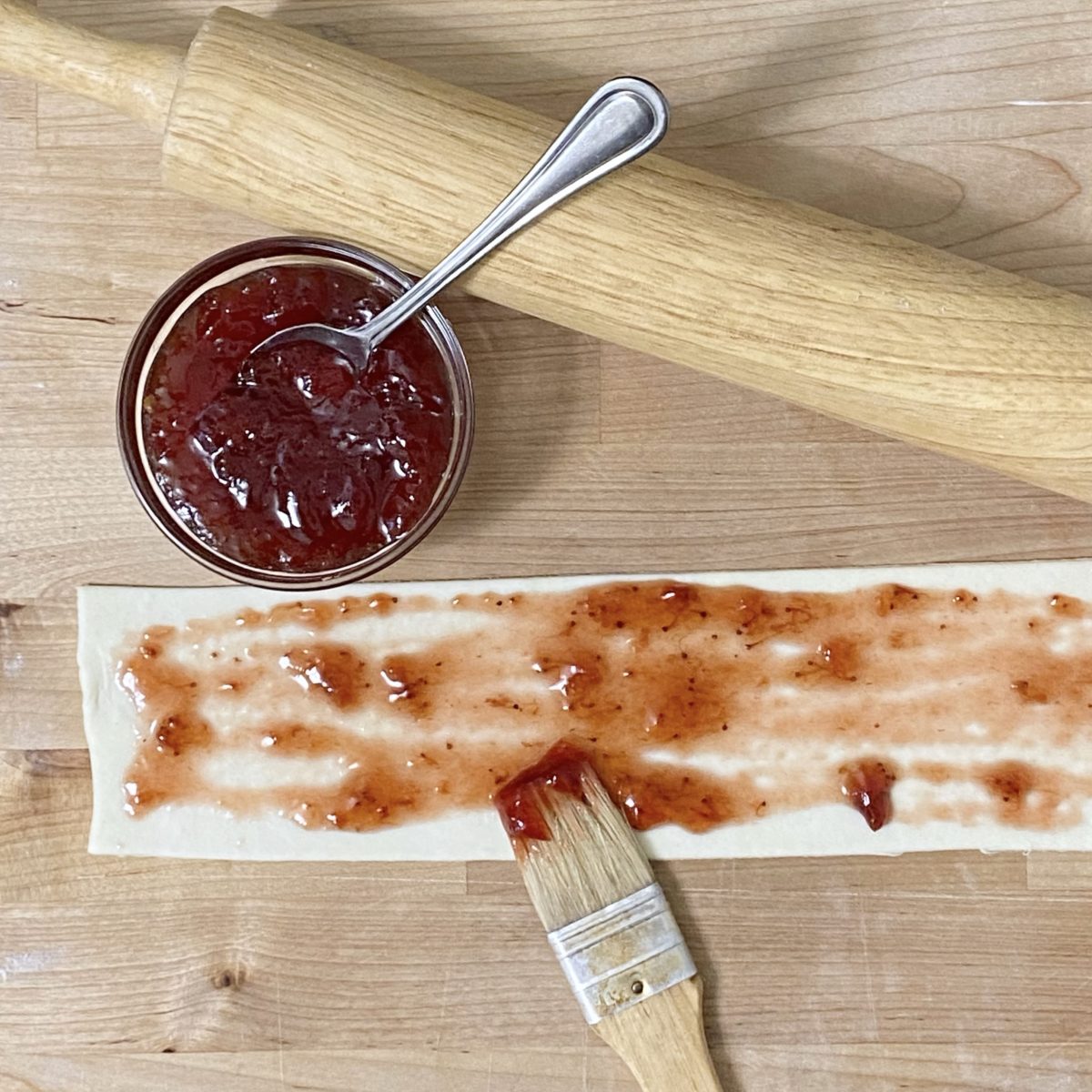 Strawberry preserves being spread on a strip of puff pastry dough with a pastry brush.