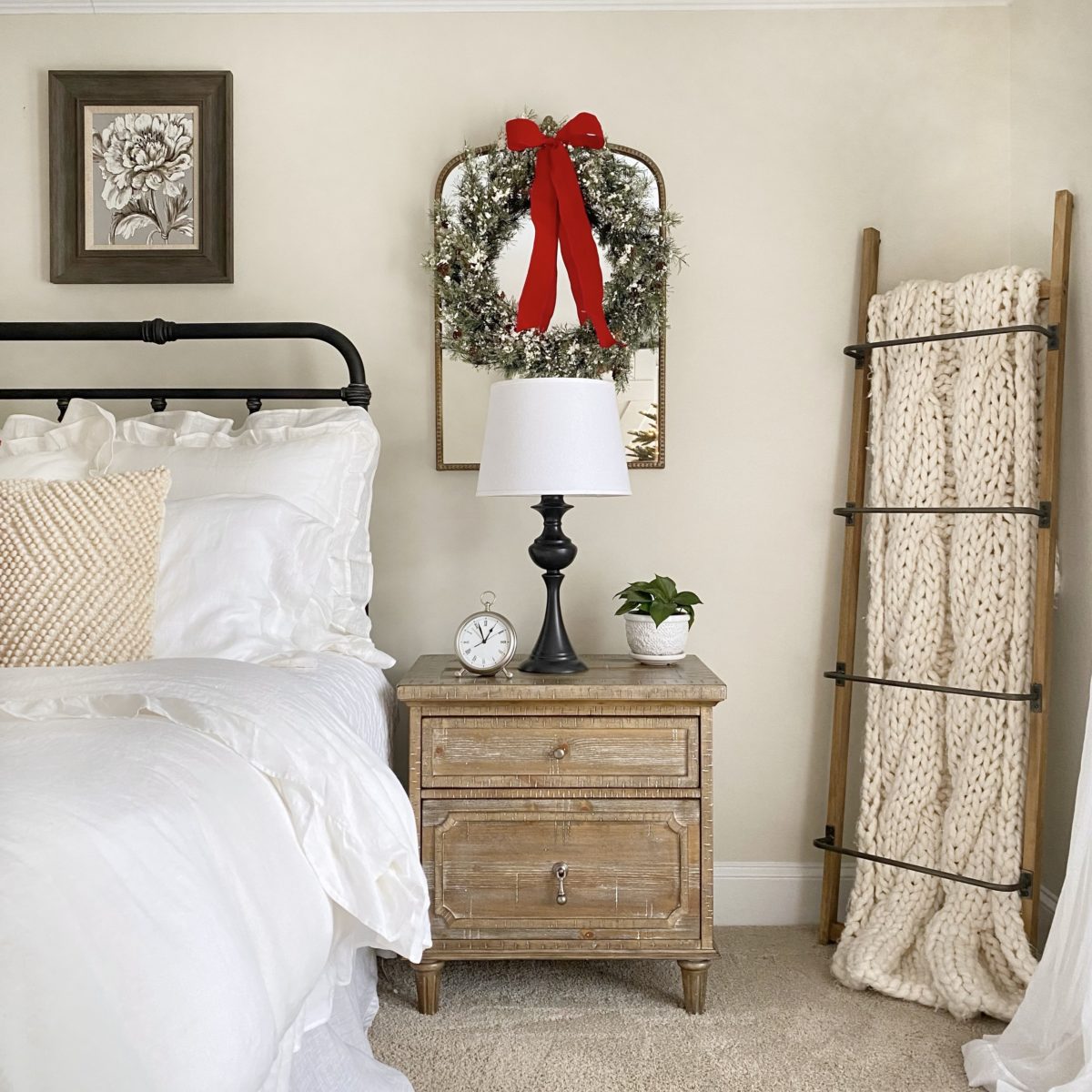The opposite side of the bed with a mirror on the wall and wreath hanging from it with a red ribbon. In the corner is a blanket ladder with a cozy knit blanket on it.