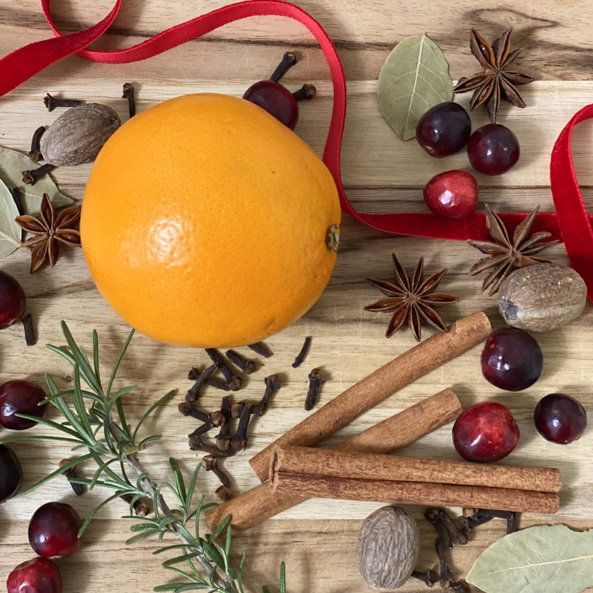 Ingredients for a DIY simmer pot gift including an orange, cinnamon sticks, cranberries, cloves, nutmeg, bay leaves, rosemary, and red ribbon.