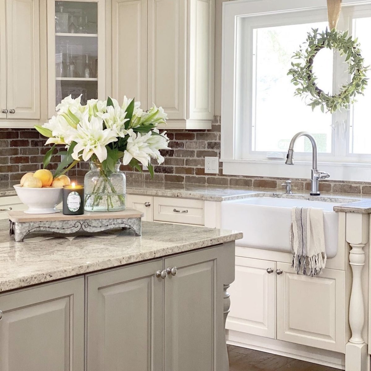 A view of the kitchen with the sink and brick backsplash in the background. There are flowers, fruit, and a candle lit on the kitchen island.