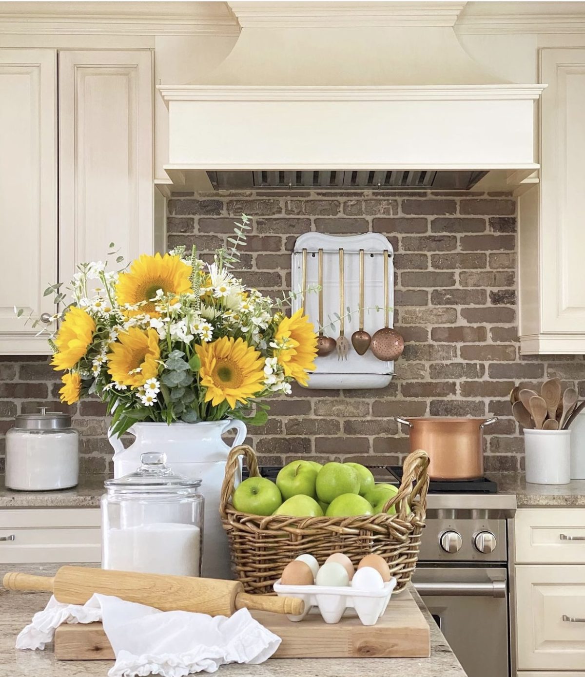Kitchen view with brick behind the range going up to the vent hood. In the foreground are sunflowers, a basket of apples, fresh eggs, and a rolling pin.