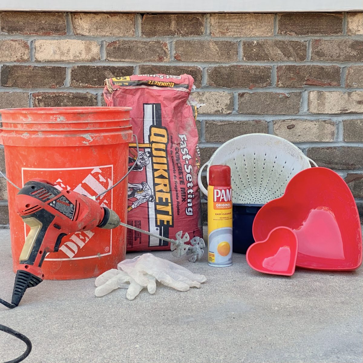 Materials needed to make DIY concrete heart dishes including quick-crete, a bucket, gloves, cooking spray, and heart shaped bowls.