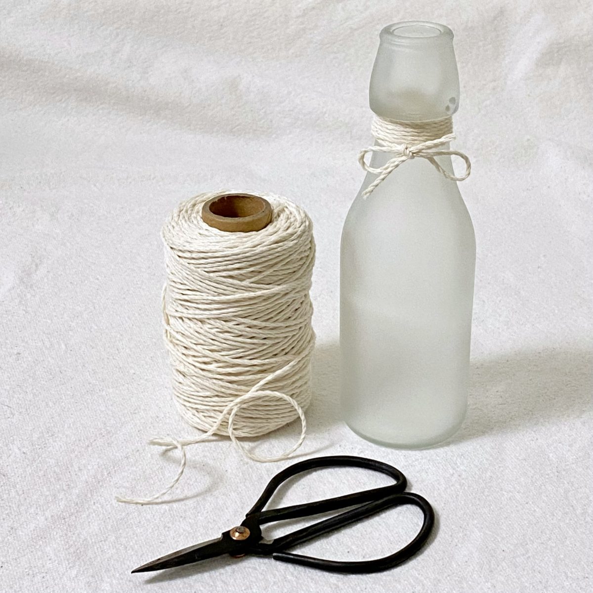 Frosted glass bottle next to a spool of string and a scissor.