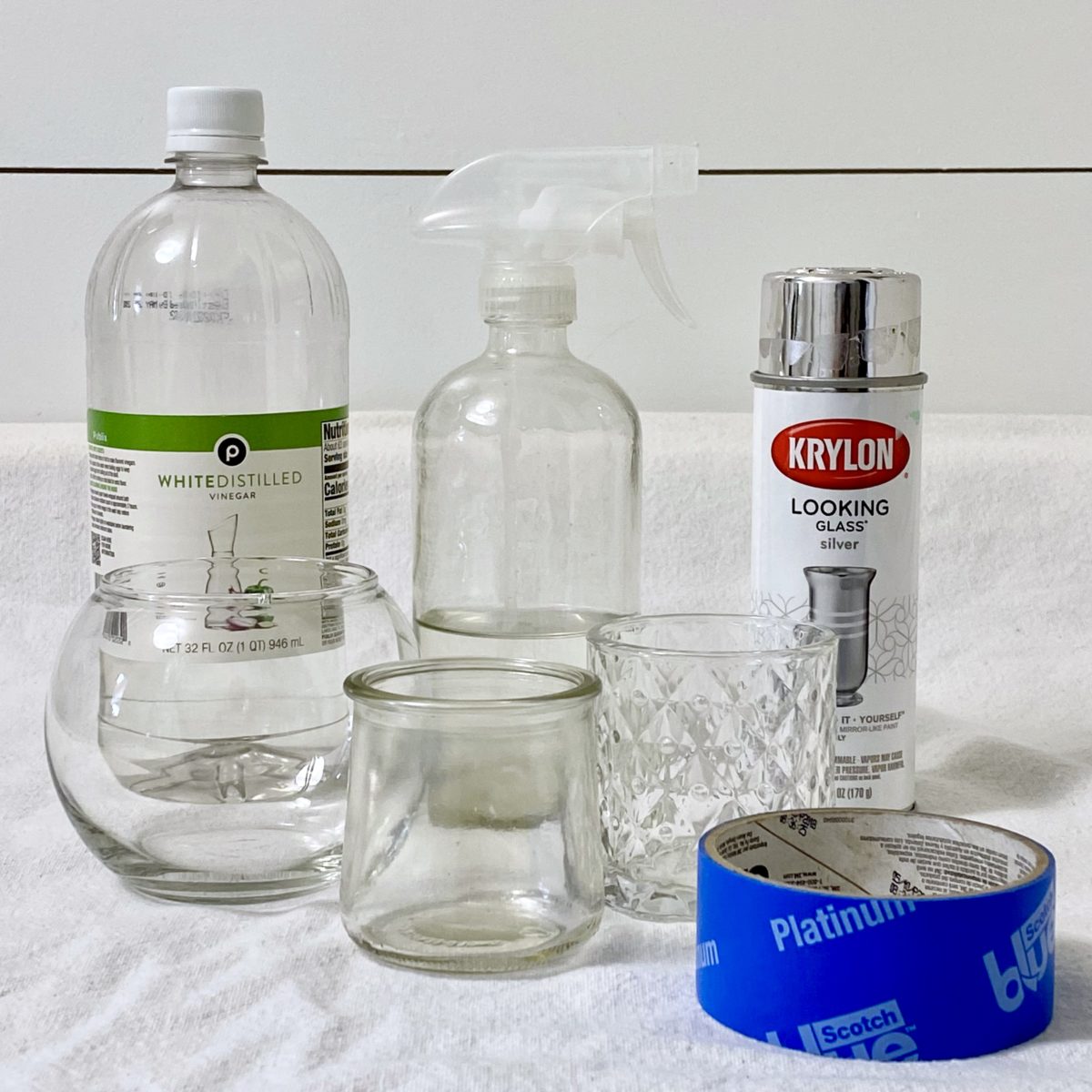 Materials needed to make DIY mercury glass candle holders including glass jars, mirror spray paint, painters tape, vinegar and water.