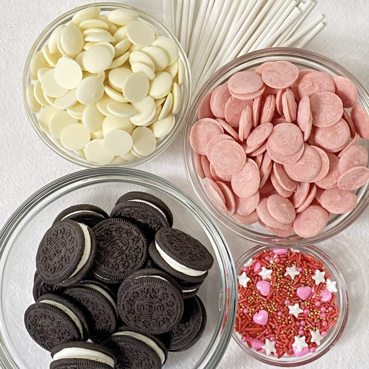 Ingredients to make Valentine's Day Oreo pops including white and pink melting wafers, Oreos, Valentine's sprinkles, and lollipop sticks.