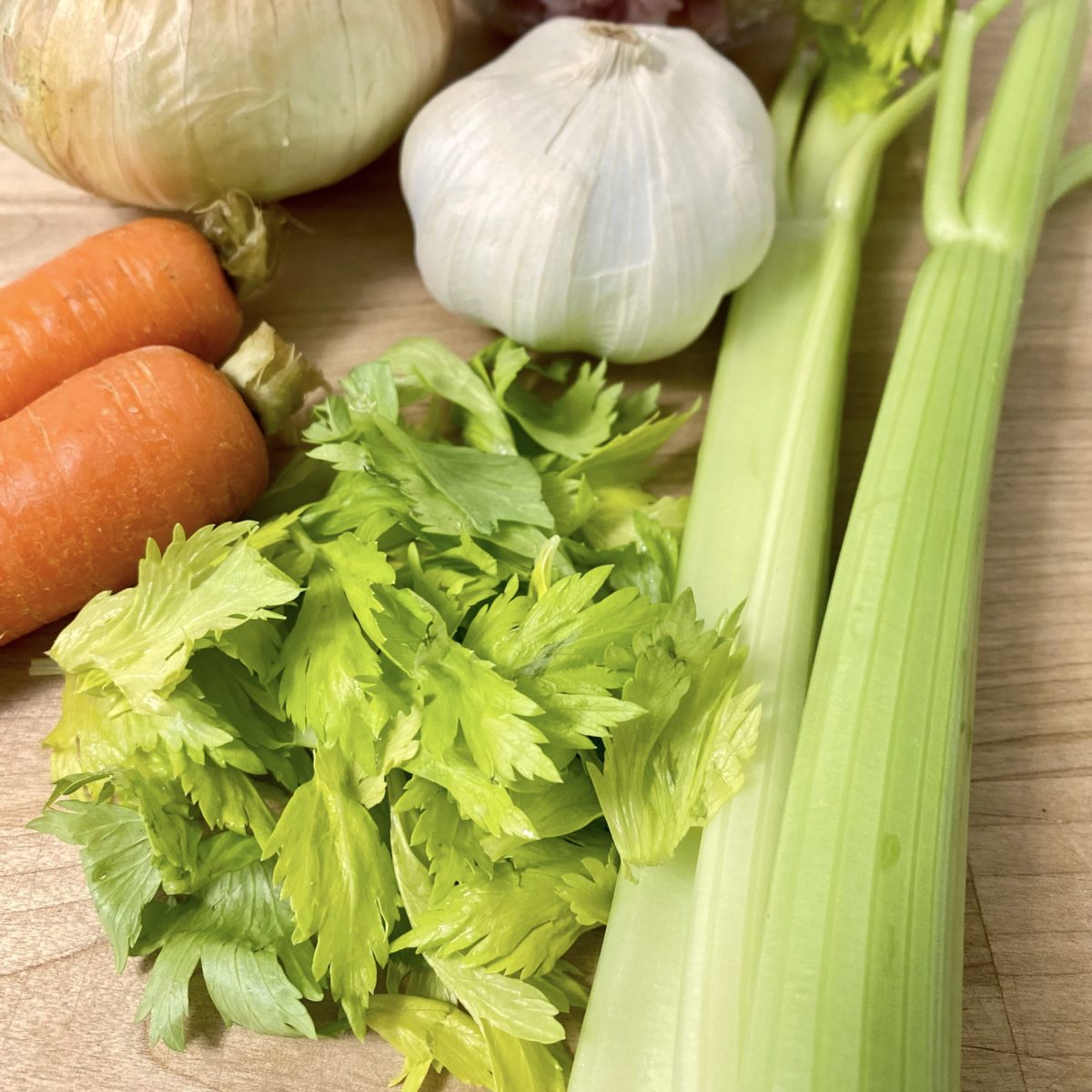 Carrots, garlic, onion and celery stalks on a cutting board with a big pile of celery leaves.