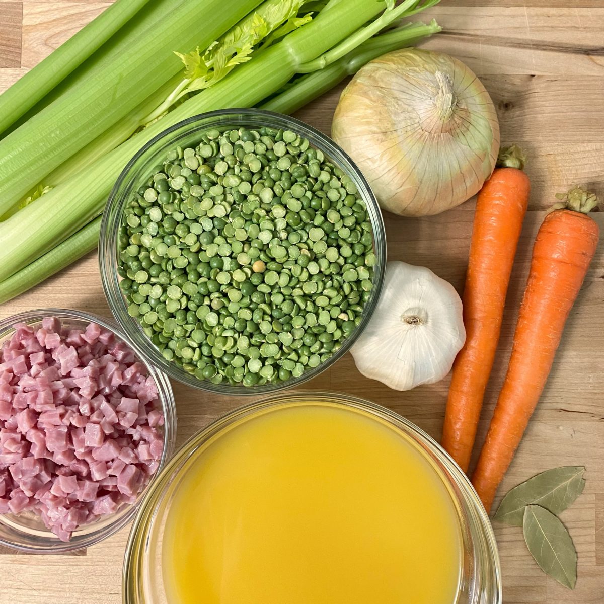 Dried split peas, chicken broth, cubed ham in bowls with celery, carrots, onion, and garlic on a cutting board.