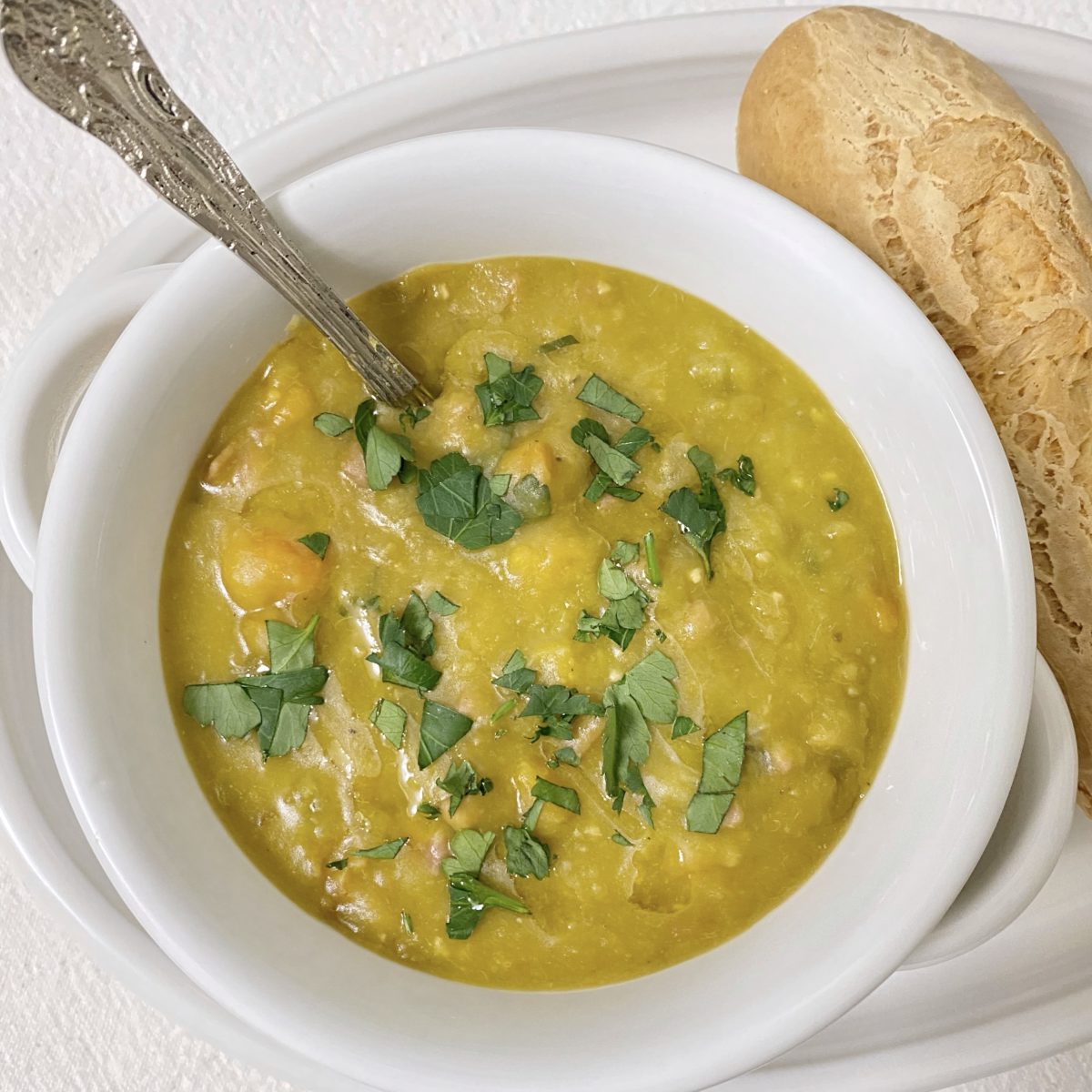 My mama's homemade split pea soup in a bowl with a spoon and a piece of bread next to it.