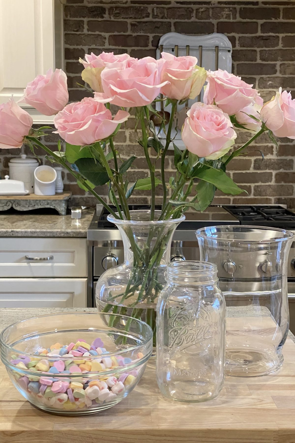 A vase, a large jar, heart candies in a bowl and pink roses for making a sweet Valentine's Day floral arrangement.