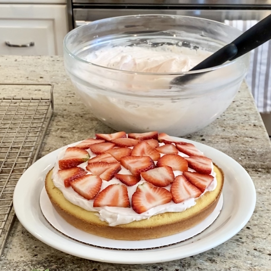 Layer of fresh strawberries on the frosting of the bottom cake. A bowl of frosting is behind it.