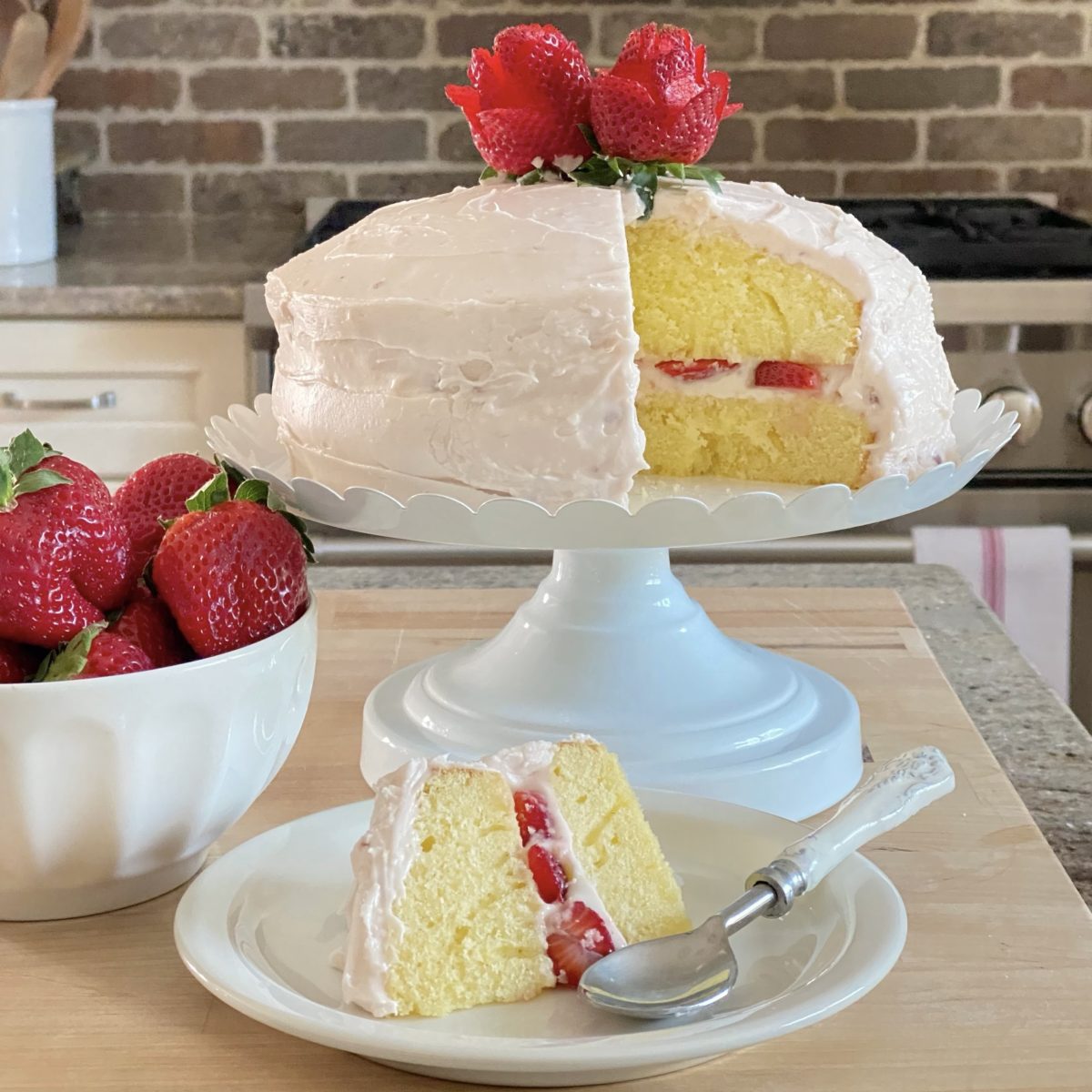 The best vanilla strawberry cake on a cake stand with a slice out of it on a plate below and a bowl of strawberries beside them.