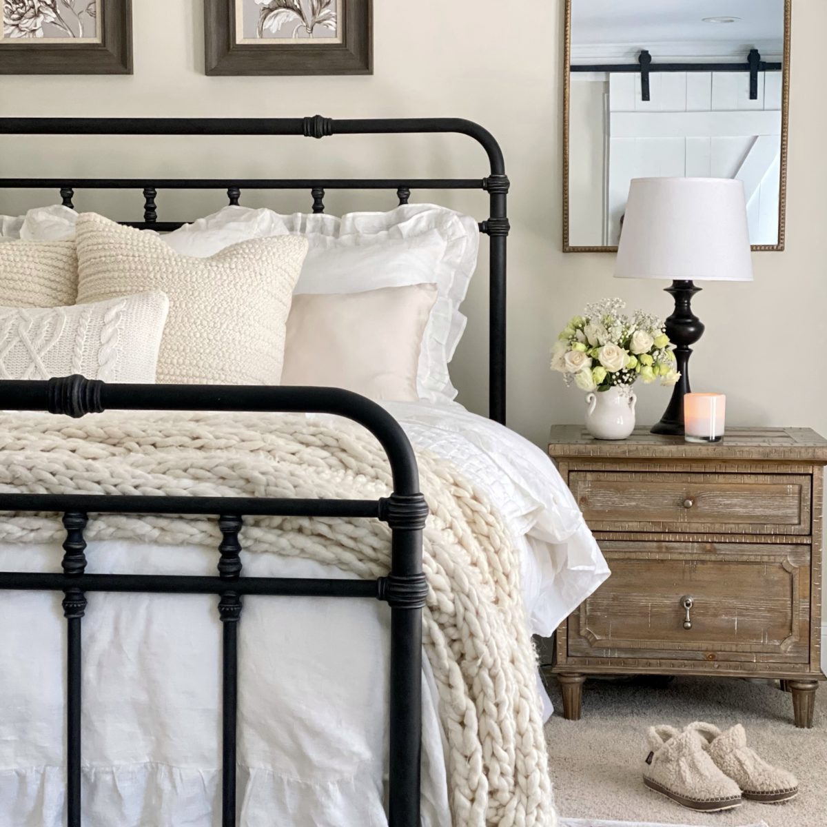 Iron bed with layered winter white bedding on it. There is a nightstand next to the bed with white flowers in a vase, a lamp, and a candle.