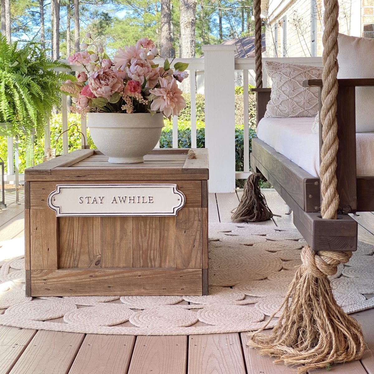 Side view of the porch swing on the front porch with a wood chest in front of it for a coffee table. On the coffee table is a pink floral arrangement.