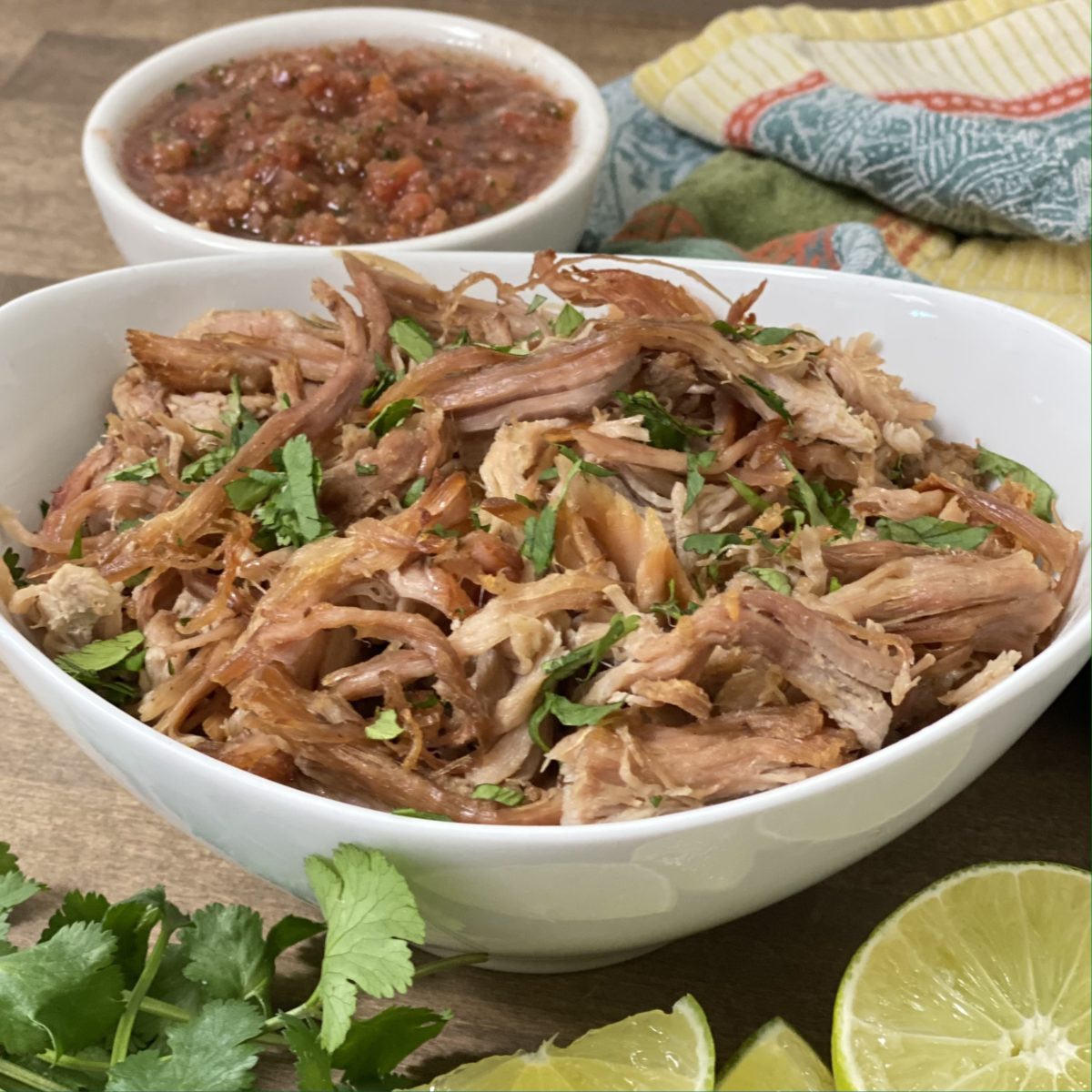 Easy slow cooker carnitas in a white bowl with lime and cilantro in the foreground and salsa in the background.