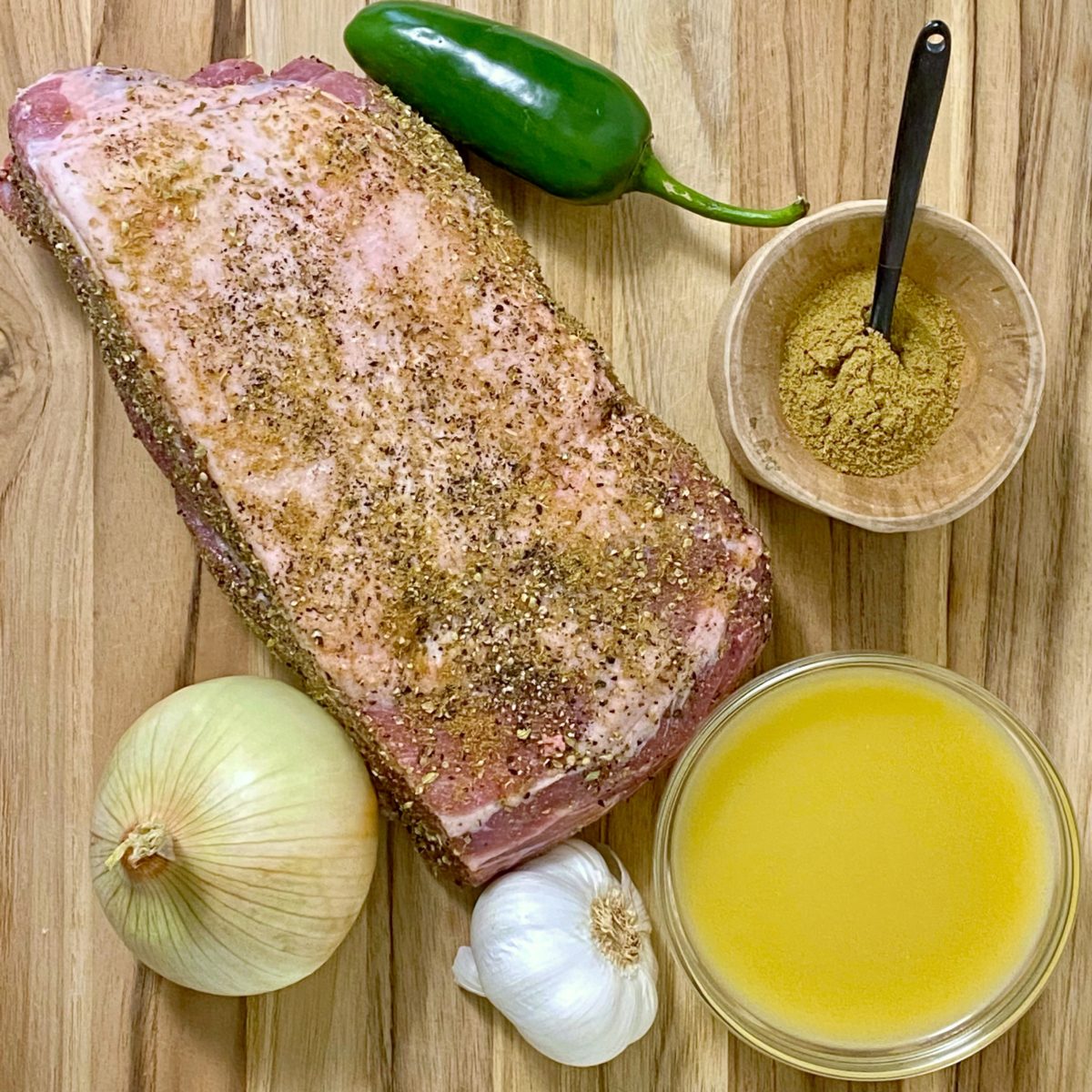 Ingredients to make carnitas o a wood board including pork butt roast, chicken broth, onion, garlic, jalapeño, and cumin.