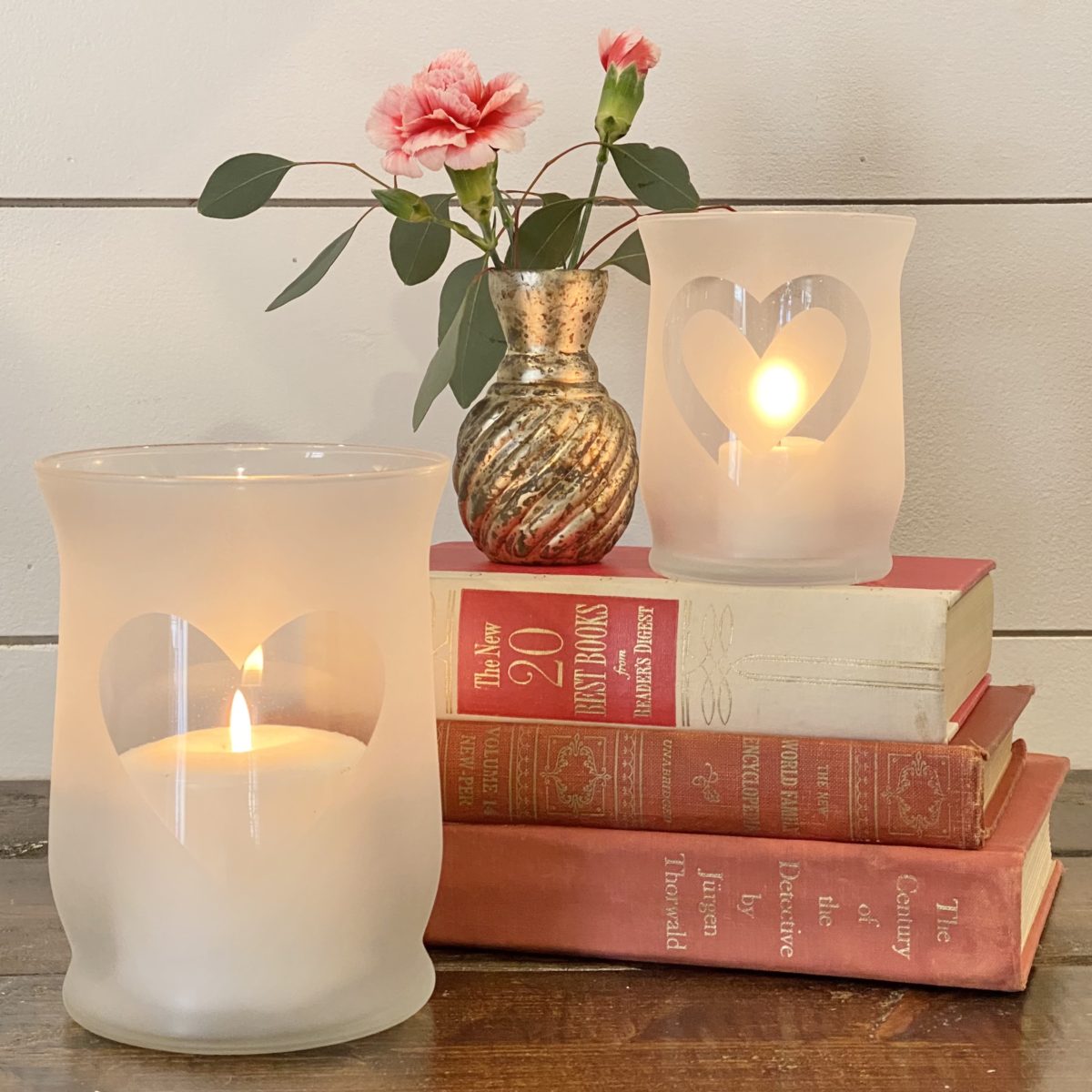 Frosted glass heart candle holders with candles lit in them styled with red, white, and pink books and a small vase with flowers.