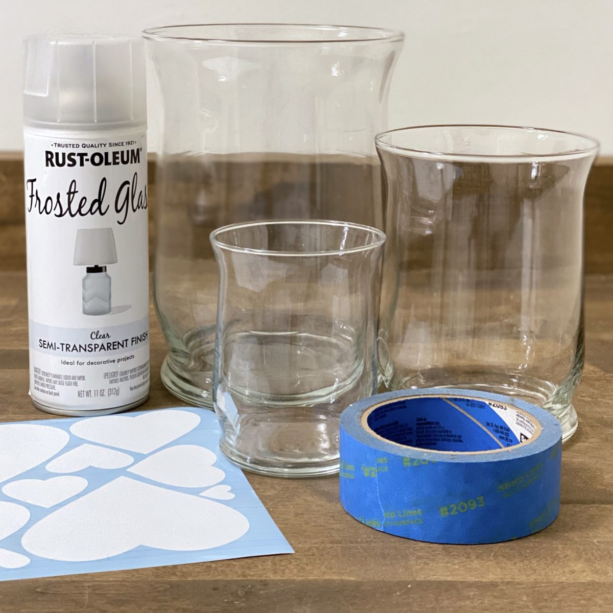 Materials needed to make frosted glass Valentine's Day candle holders out of glass hurricanes using painter's tape, heart decals, and frosted glass spray paint.