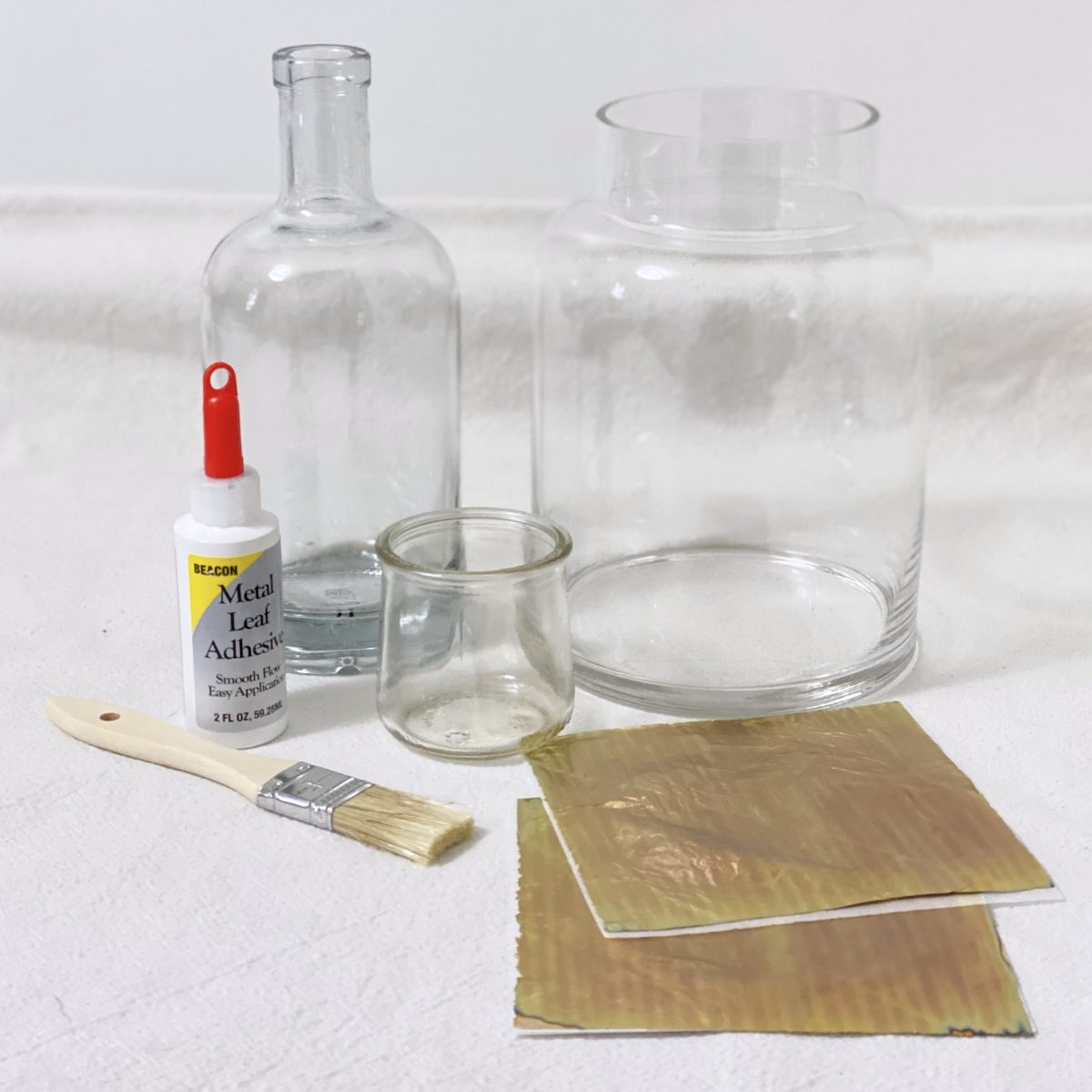 Items needed to make a DIY gold leaf vase including a glass vase, gold leaf sheets, adhesive, and a paint brush.