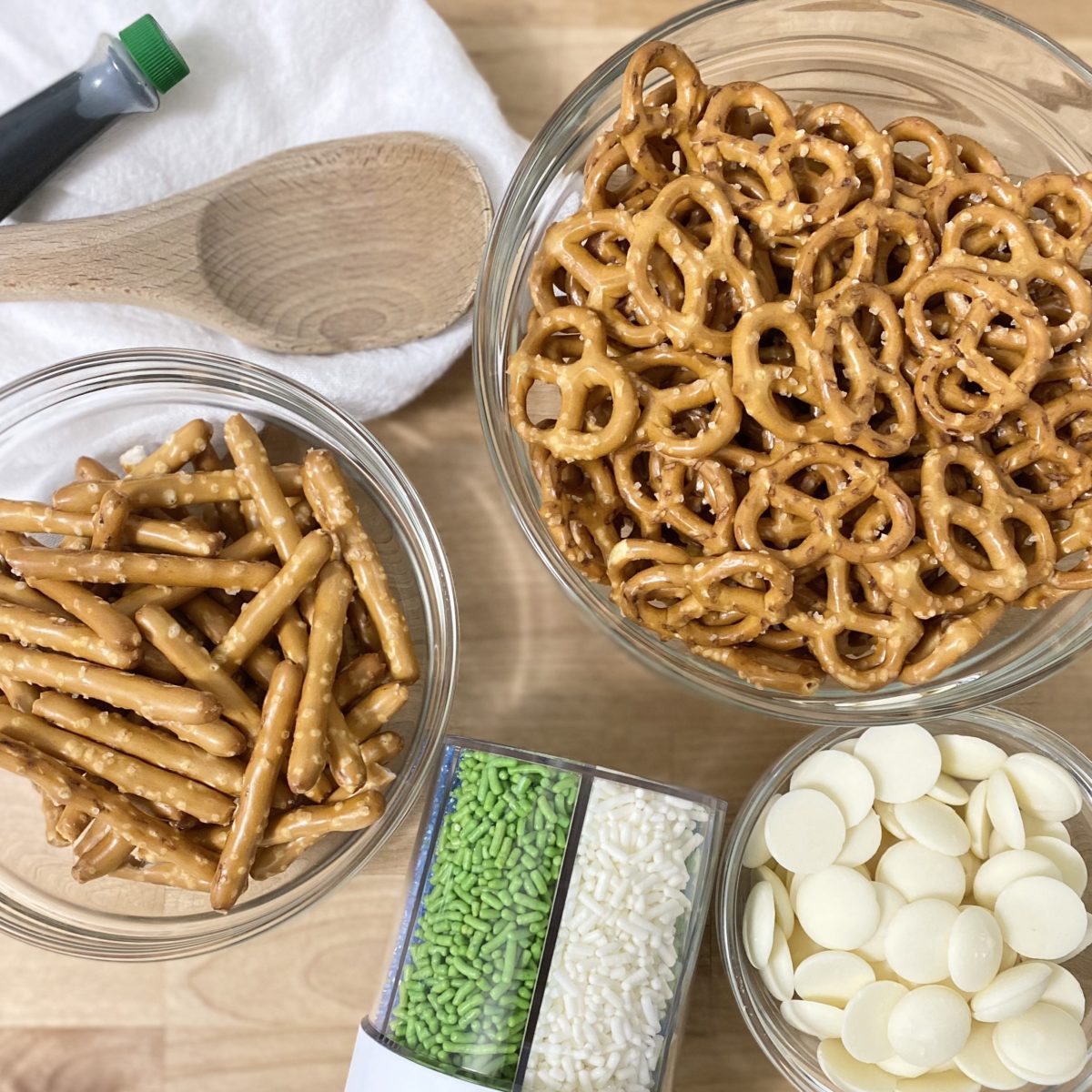 Ingredients to make pretzel shamrocks including pretzels, melting chocolate wafers, green food coloring, and sprinkles.
