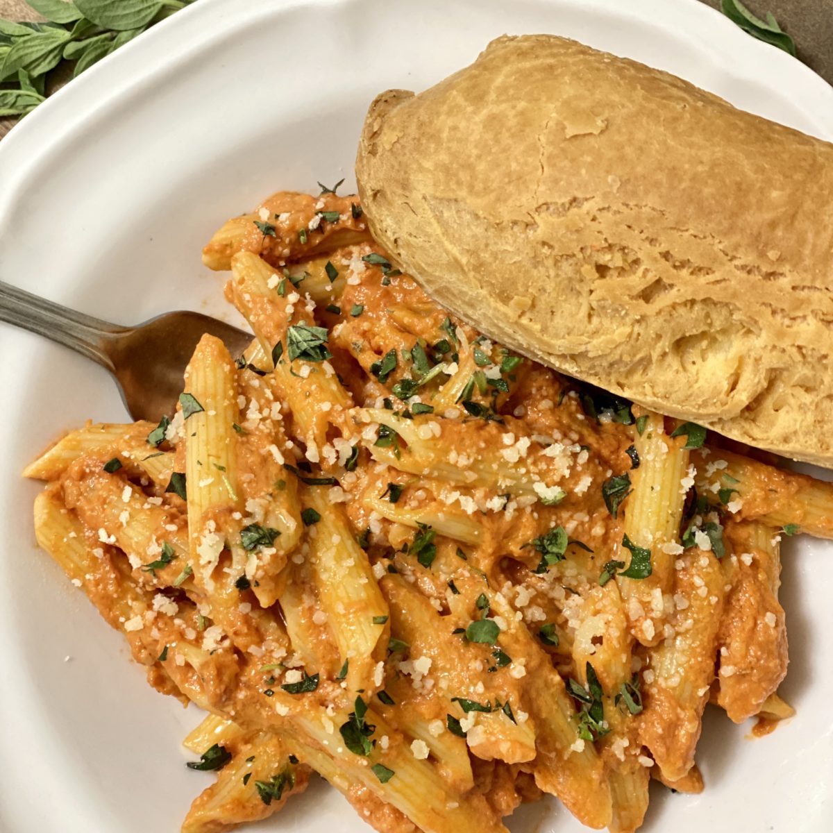 Vodka penne in a white bowl garnished with chopped fresh oregano and Parmesan cheese. A baguette is on the side.
