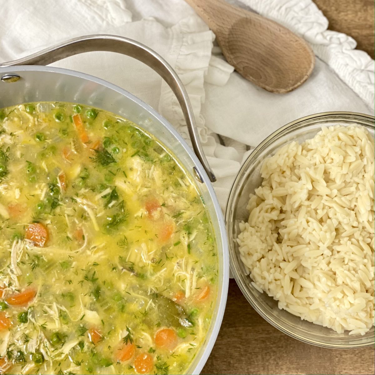 Chicken and orzo soup with lemon and dill in a pot with a bowl of orzo and a wooden spoon next to it.
