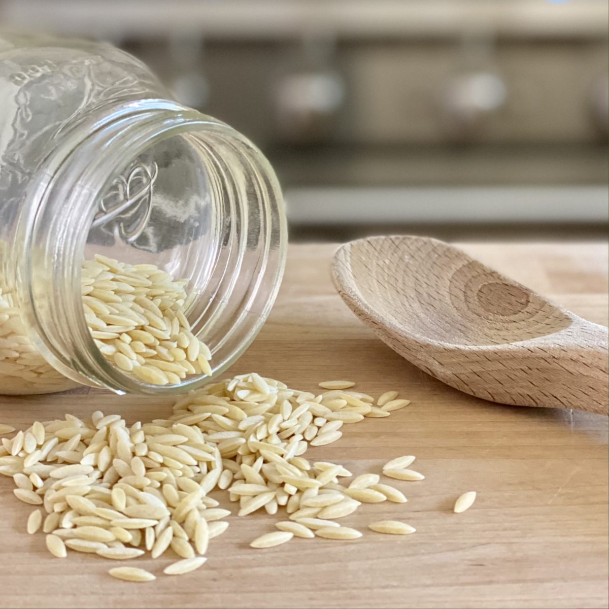 Orzo spilling out of a glass jar with a wooden spoon next to it.