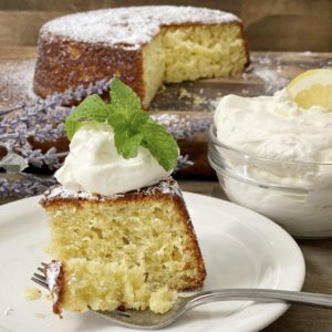 A slice of lemon and lavender cake on a white plate with a dollop of lemon whipped cream and a mint spring on top. In the background is the whole cake.