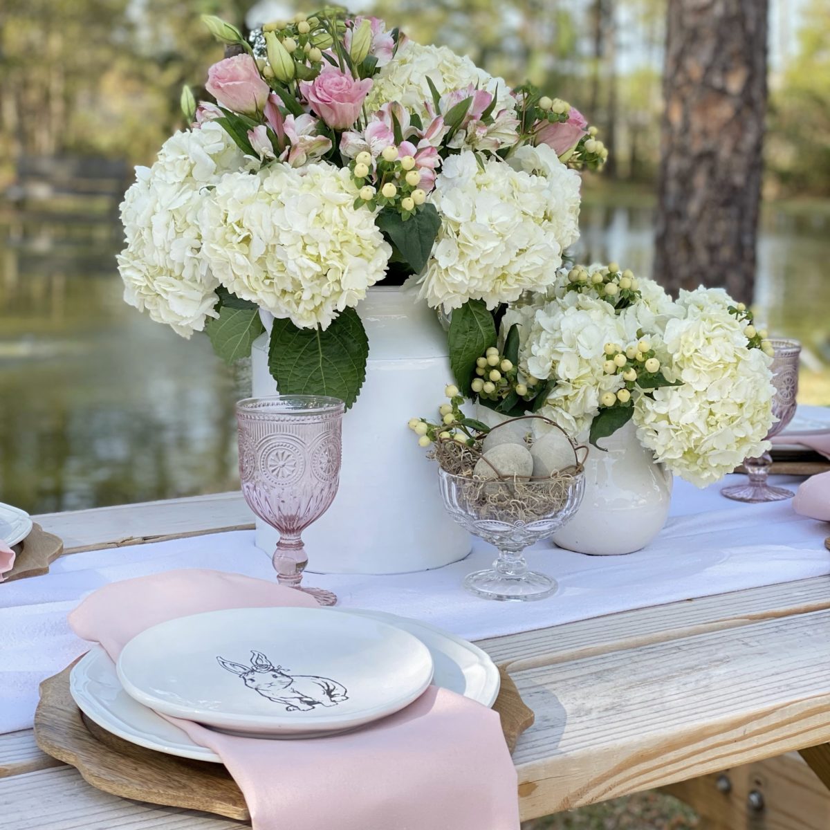 A simple spring table set with white dishes and pink accents on a picnic table by a pond,