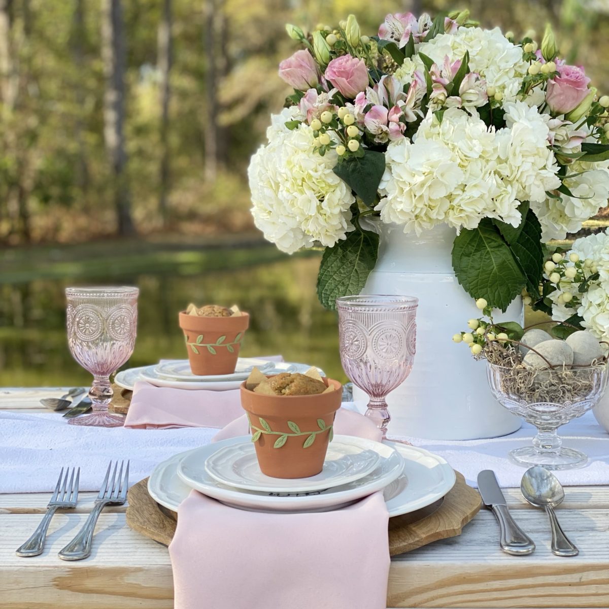 A simple spring table set with white dishes and pink accents on a picnic table by a pond.