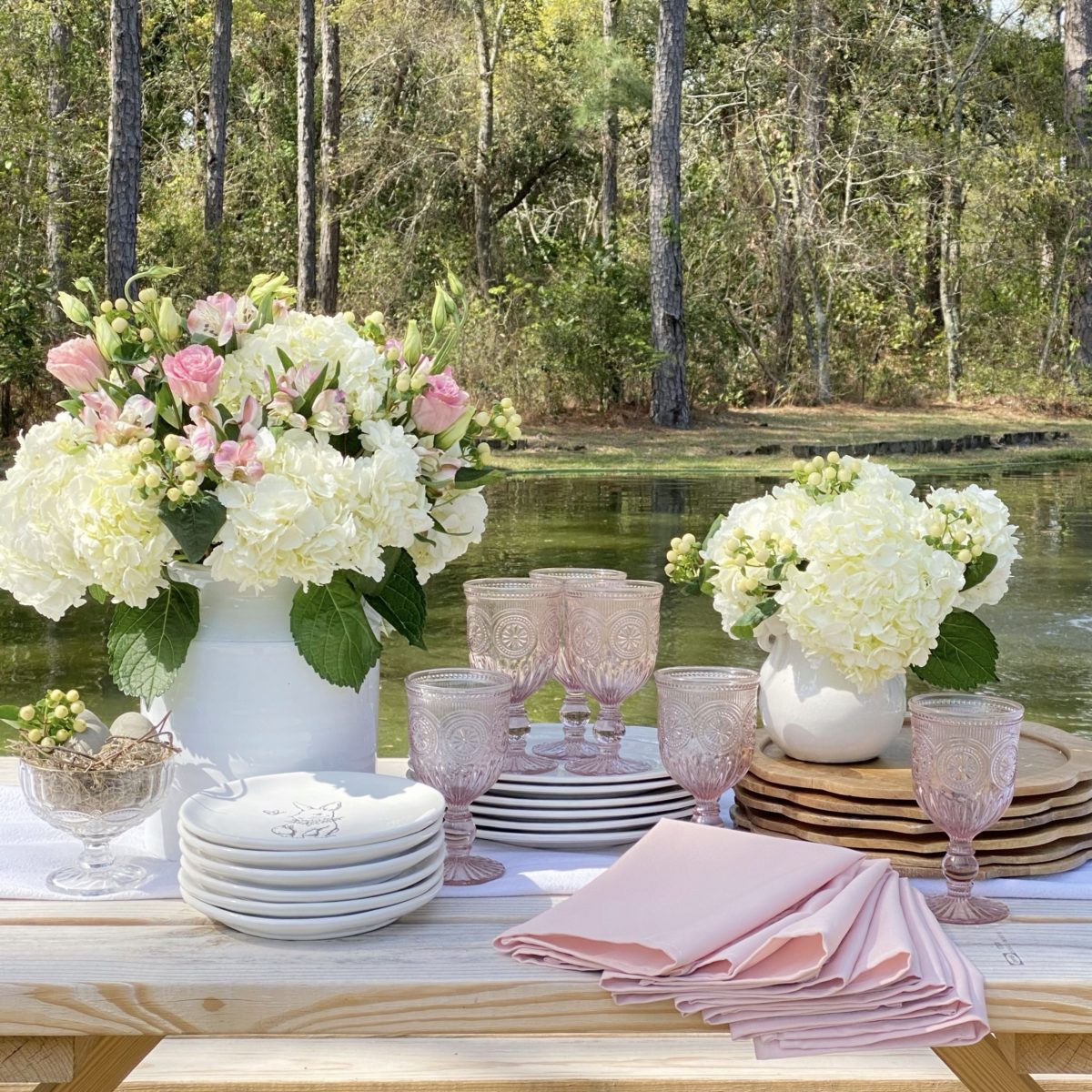 Wood chargers, white plates, pink napkins and glass with spring florals ready to be set on the picnic table by the pond., 