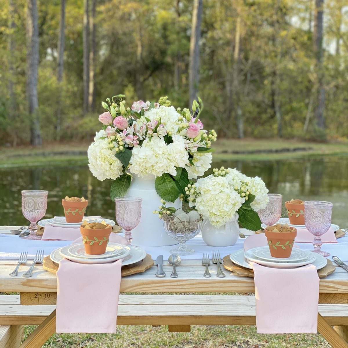 A simple spring table set with white dishes and pink accents by a pond.