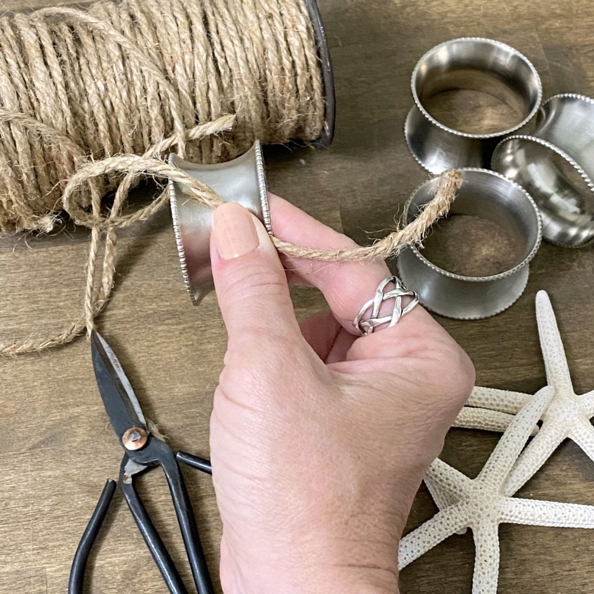 Starting to wrap twine around silver napkin rings by hold the end of the twine perpendicular with your finger leaving about a three-inch tail.