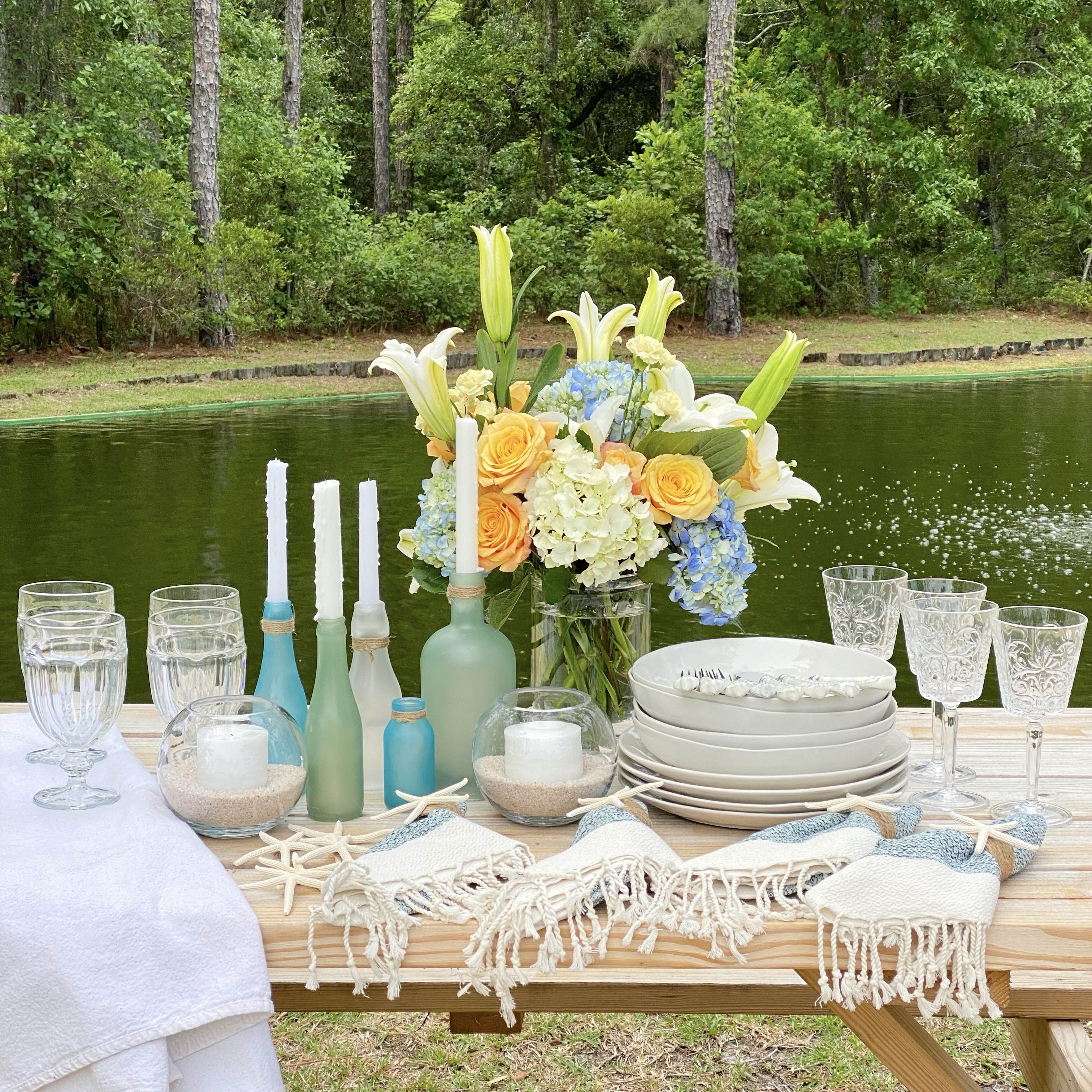 Items to be used in a simple summer tablescape set out on a picnic table by a pond.