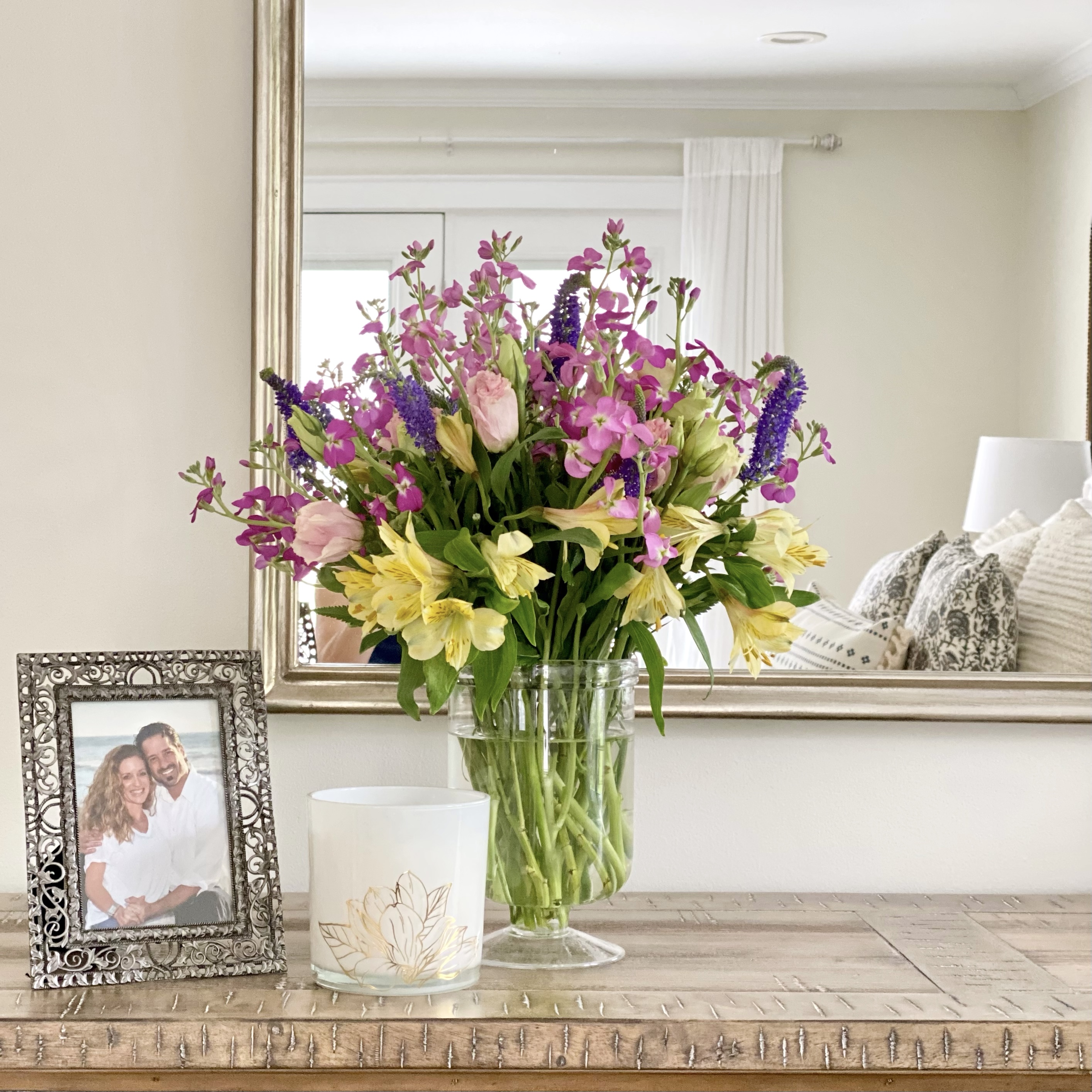 Fresh summer flowers on a dresser with a photo in a frame and a candle a mirror is in the background.