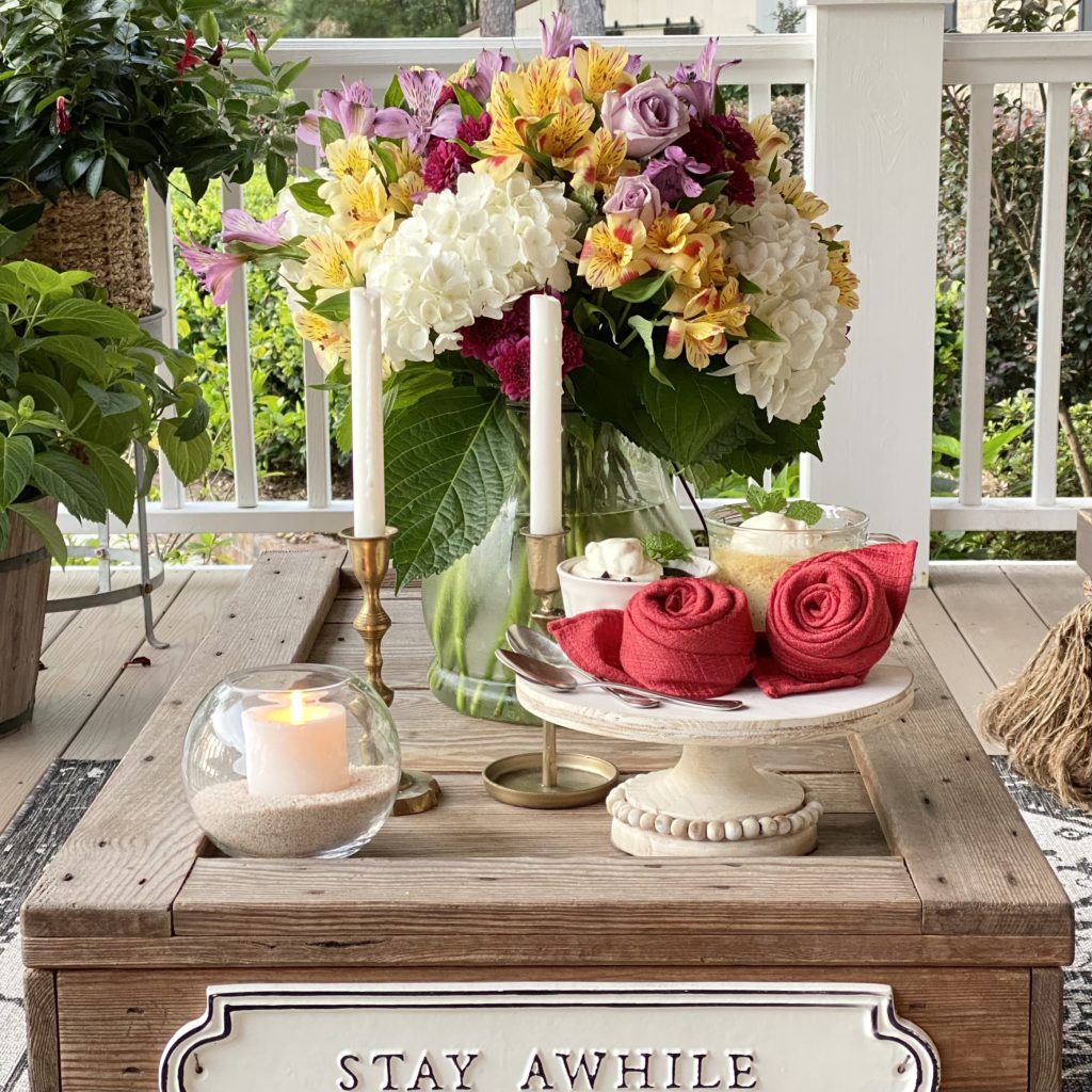 Red cloth napkins folded into roses on a pedestal tray with candles and flowers on top of a coffee table on a porch.