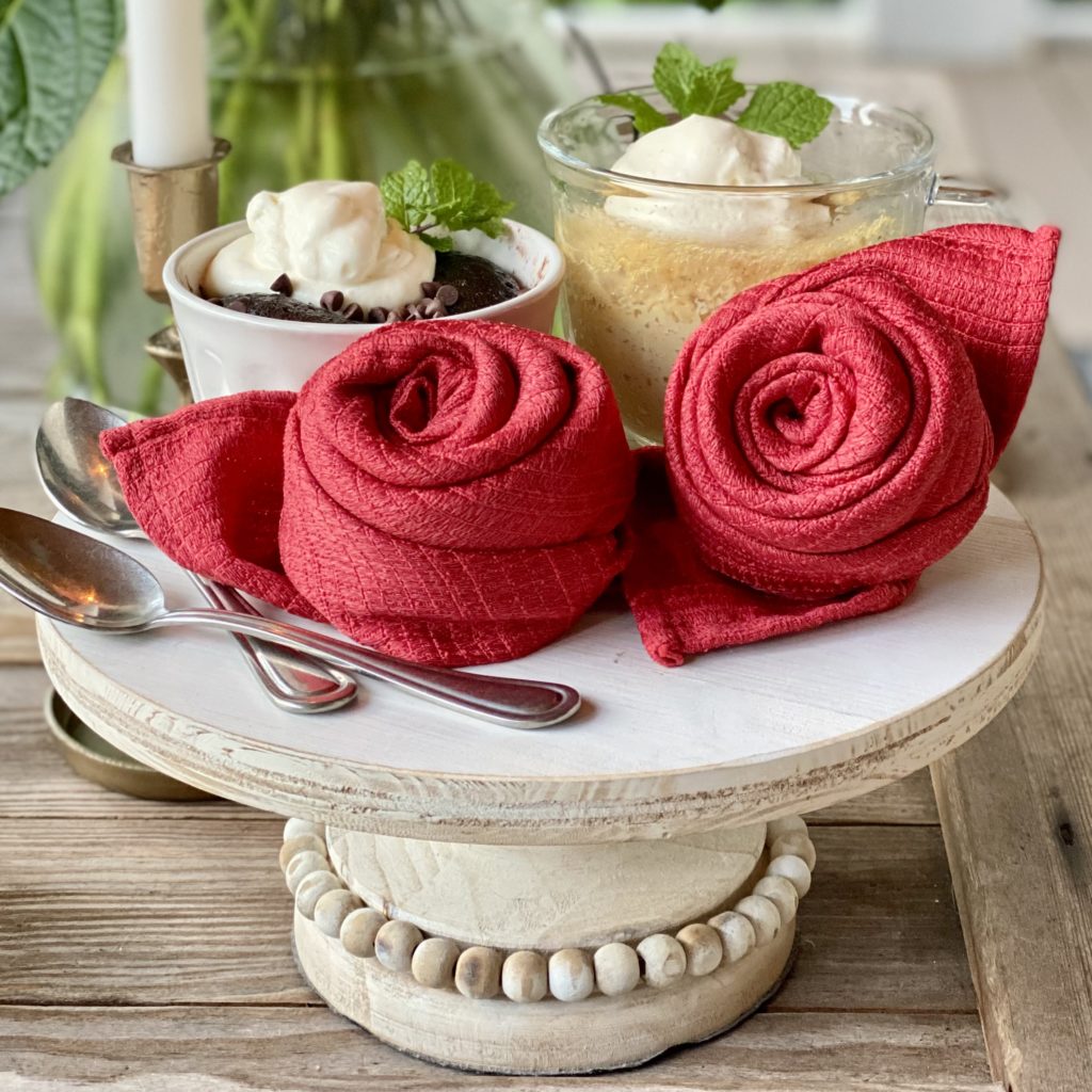 Red cloth napkins folded into roses on a raised pedestal with mug cakes in the background.