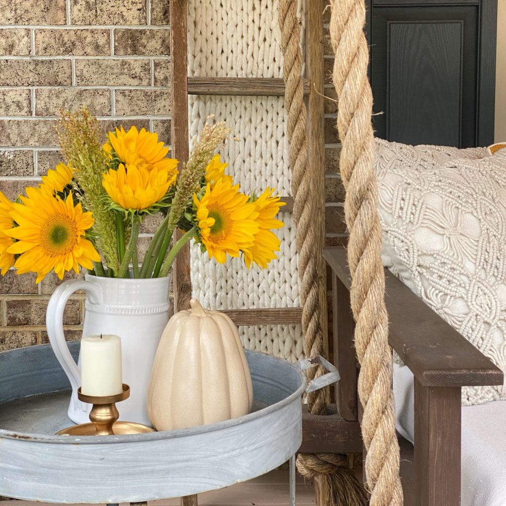 A white pitcher filled with sunflowers, a ceramic pumpkin, and candle on a side table next to the porch swing. 