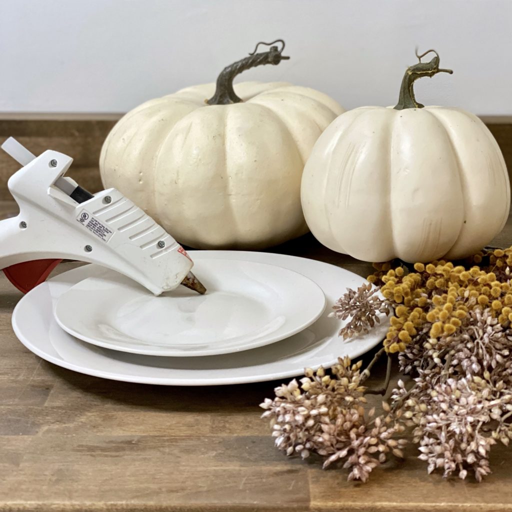 Materials needed to make a DIY tiered tray for fall including two faux pumpkins, two plates, hot glue, and faux fall stems.