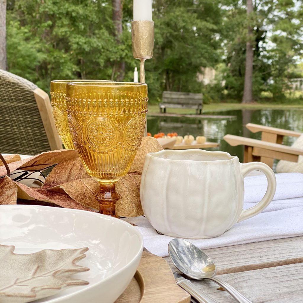 Drinkware on the table for a casual fall outdoor gathering including an amber goblet and white pumpkin ceramic mug.