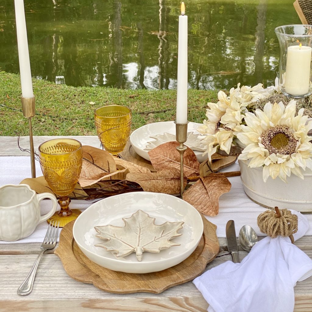 Place setting for a casual outdoor fall gathering which includes a wood charger, white shallow bowl, leaf small plate, amber goblet, and white pumpkin ceramic mug.