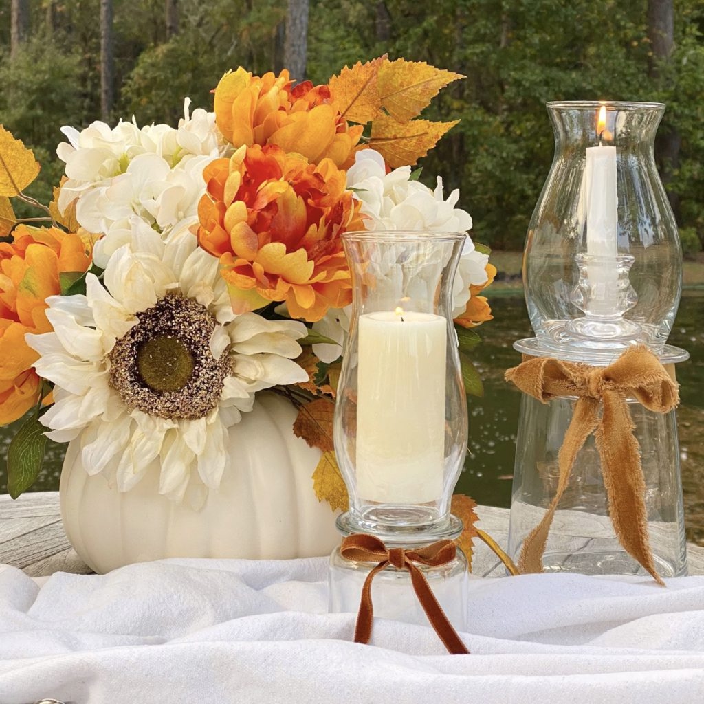 DIY glass hurricane lamps on a table outside with a faux floral arrangement in a white pumpkin.
