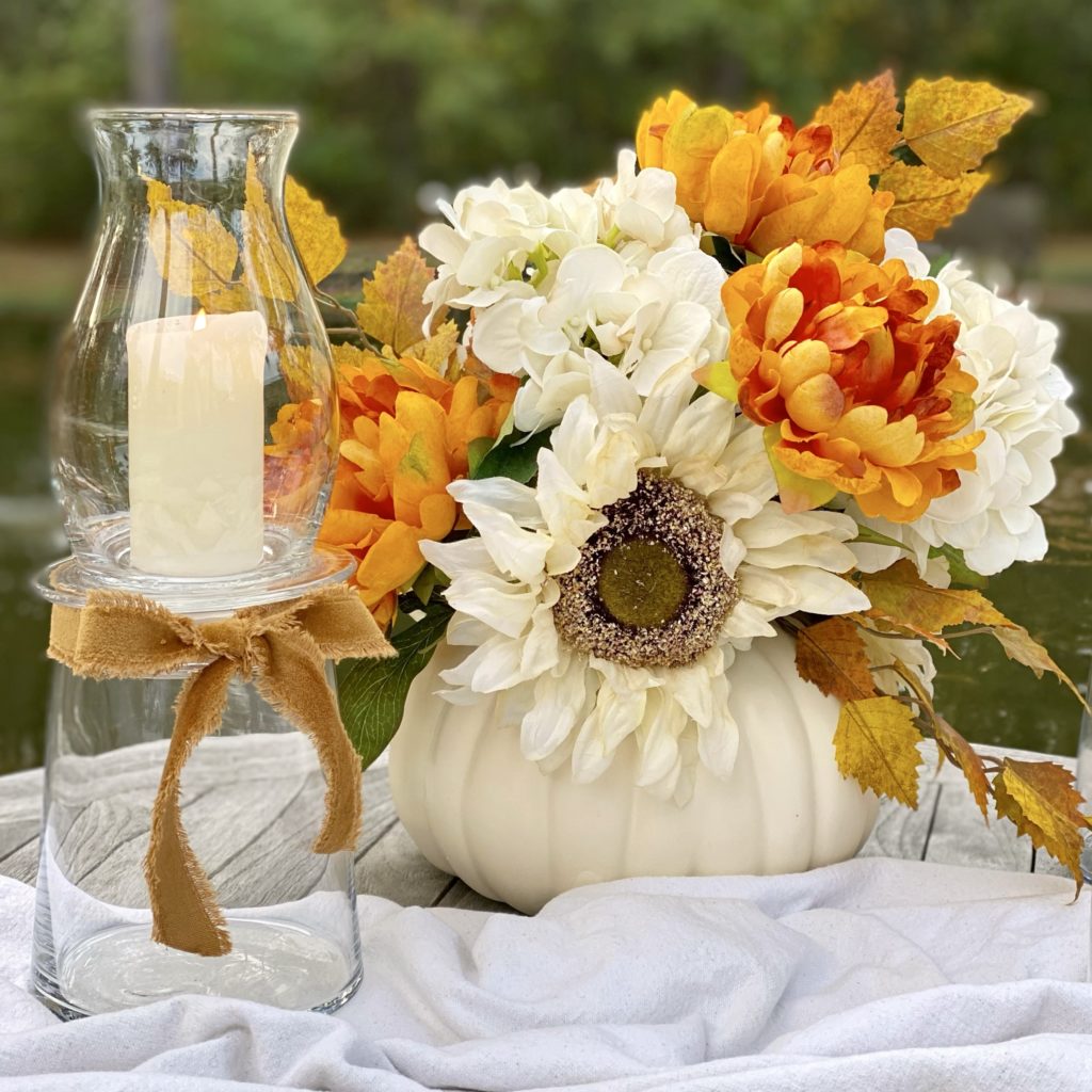 A large DIY glass hurricane lamp on a table outside with a faux floral arrangement in a white pumpkin.