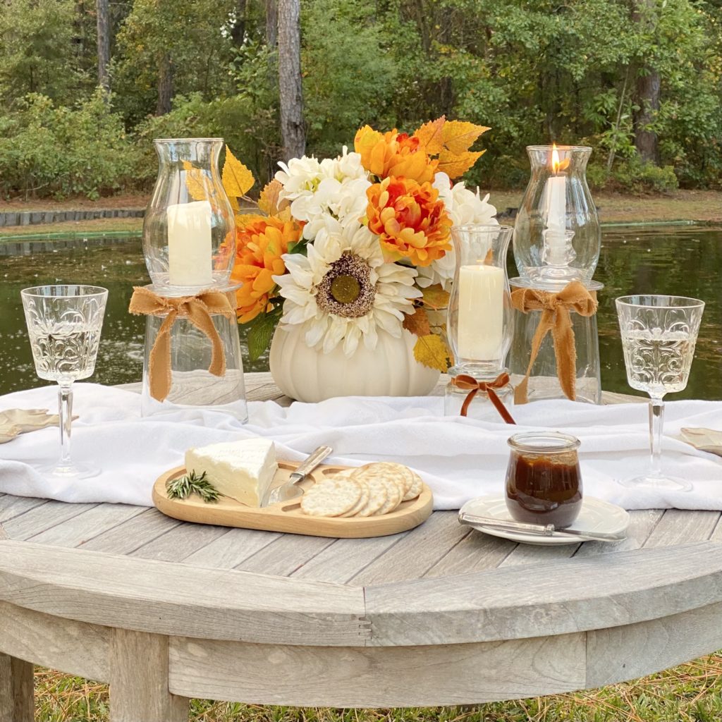 DIY glass hurricane lamps on a table by the pond with a faux fall floral arrangement, wine, and cheese.