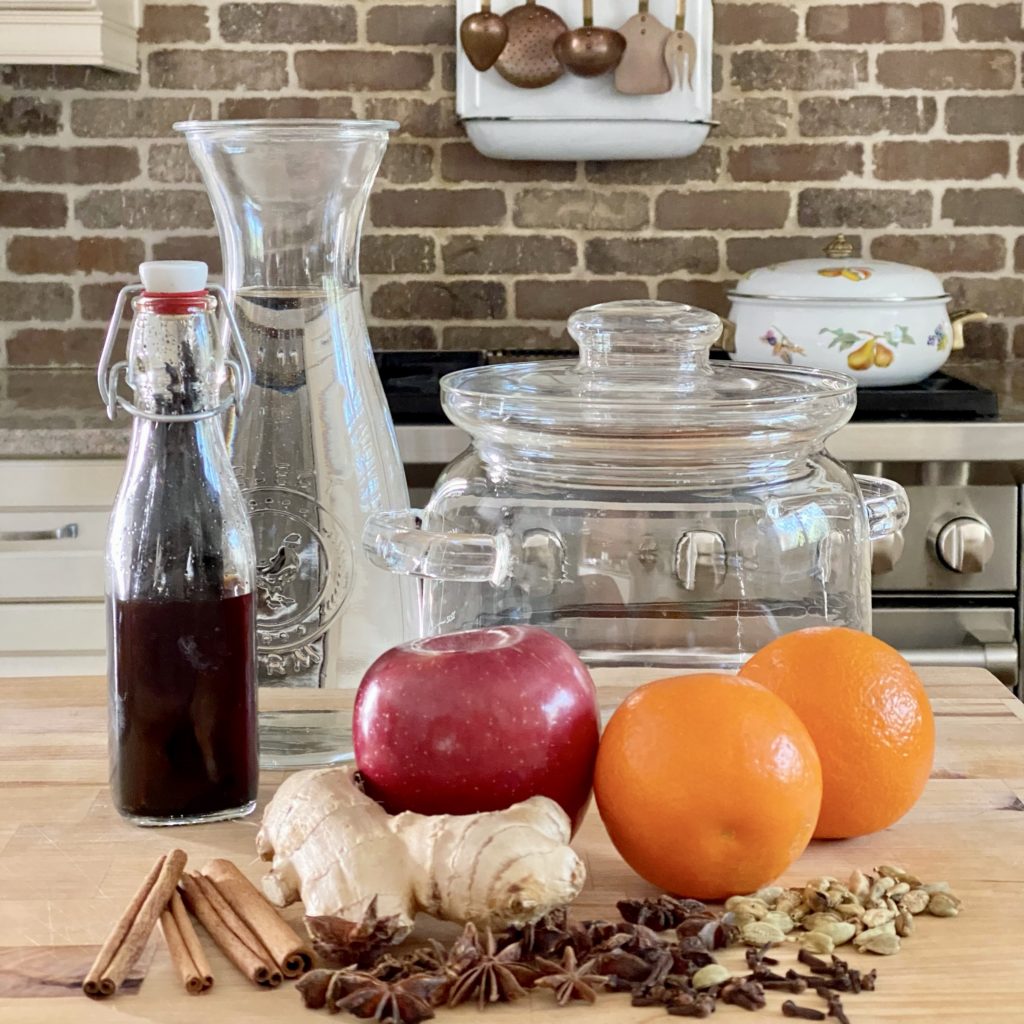 A glass simmer pot and the ingredients including apple, oranges, ginger, cinnamon, vanilla extract, and more spices to make one on the counter in the kitchen. The stove is in the background.