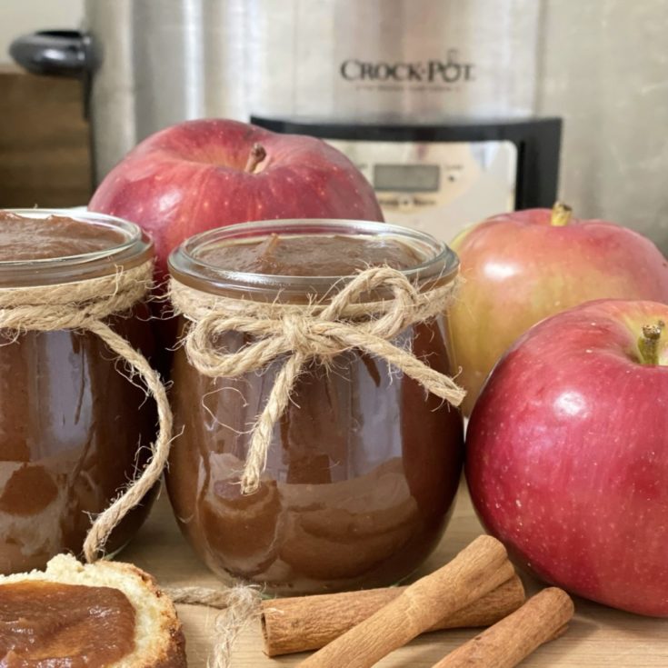 Easy crockpot apple butter in a jar with apples and cinnamon sticks in front of a crockpot.