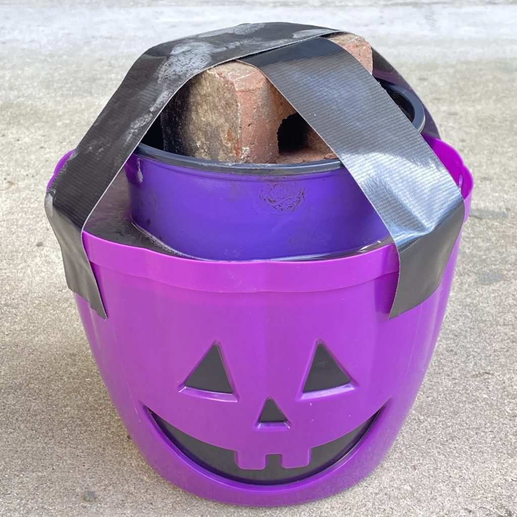 Top view of Jack O’Lantern bucket filled with concrete and smaller container inside. Inside the container is a brick holding it down in the concrete with tape over the top in an ‘X’ formation.