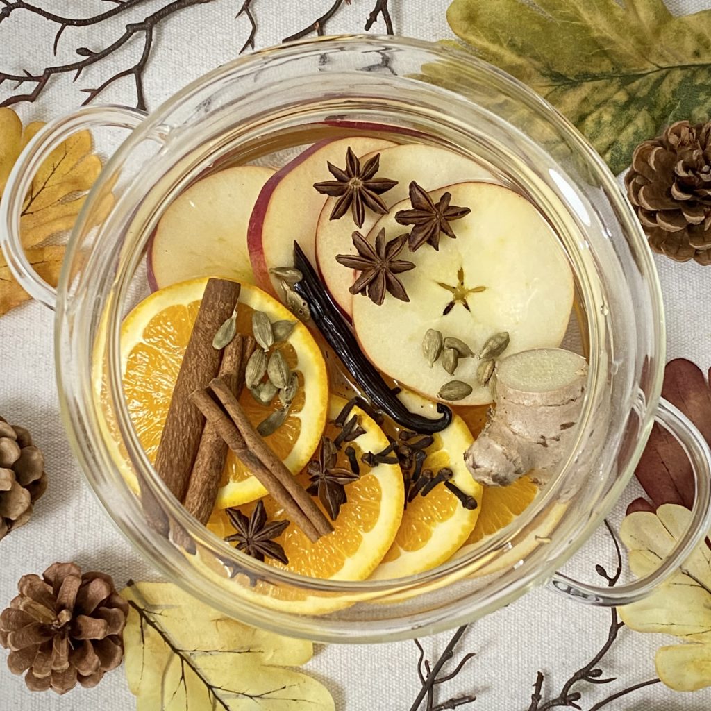 An Autumn simmer pot with apple, orange, cinnamon sticks, cardamom, star of anise and more in a glass pot.